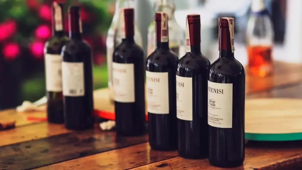 A row of dark wine bottles with white labels reading "Intenisi Rosso Puglia" lined up on a rustic wooden table, with blurred pink flowers and other bottles in the background.