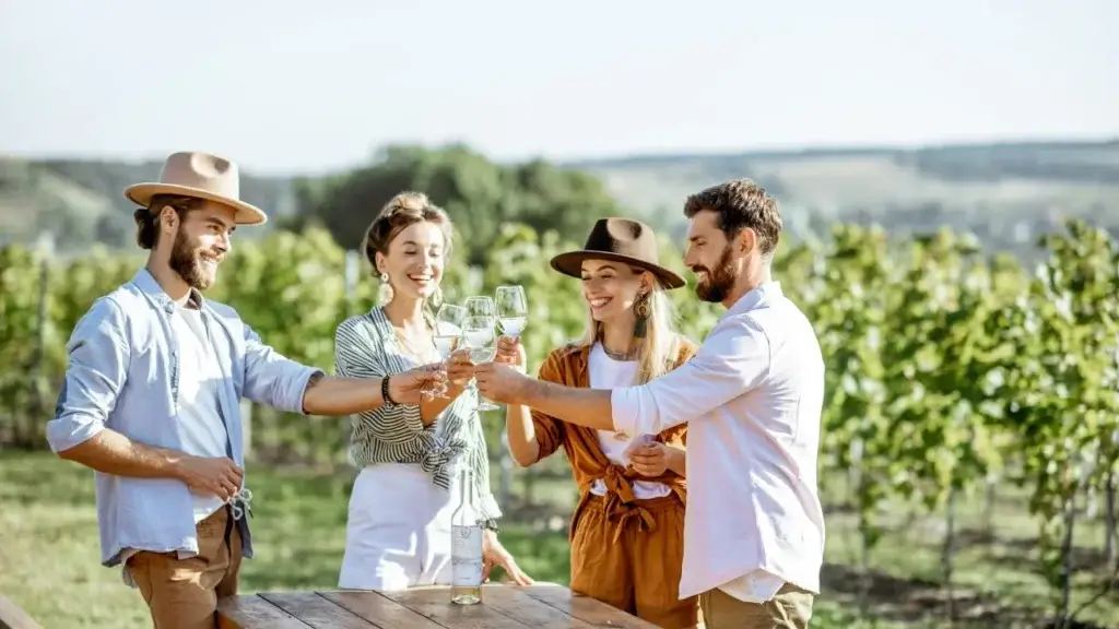 Four smiling friends wearing casual summer clothes and hats, standing around a wooden table in a sunny vineyard and clinking their wine glasses in a toast.
