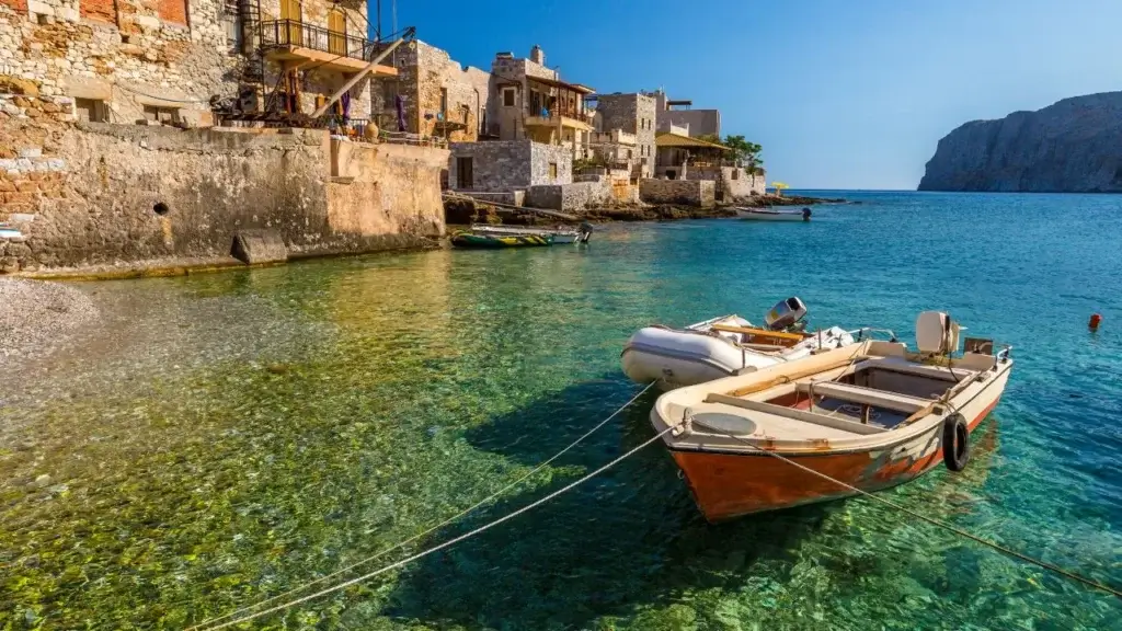 Two small boats, one motorboat with an orange hull and one inflatable dinghy, tied up in clear, shallow water next to a row of traditional stone waterfront buildings.
