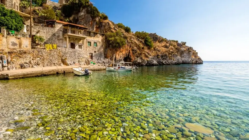 A scenic coastal cove with crystal clear, shallow water revealing a rocky seabed, featuring small boats moored near traditional stone houses built into a cliffside.