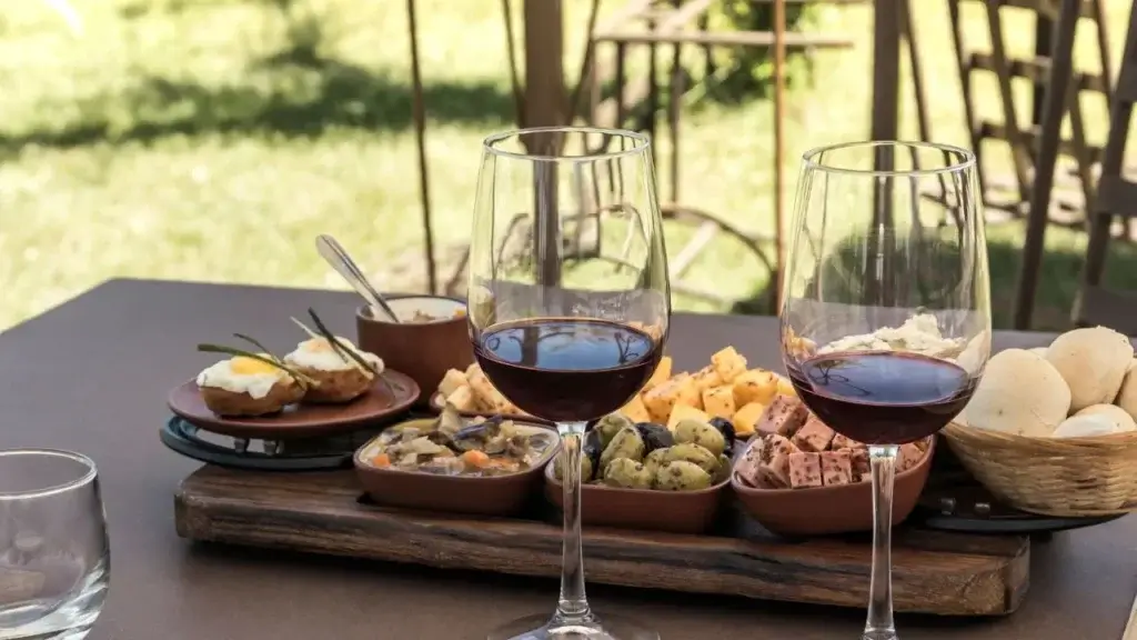 An outdoor dining table featuring two glasses of red wine and a wooden serving board filled with small clay dishes containing assorted tapas, including olives, cheese, meats, and breads.
