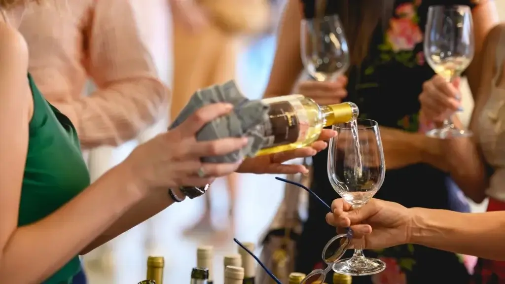 A close-up of a person wrapping a cloth napkin around a wine bottle as they pour white wine into a glass held by another person at a social gathering.
