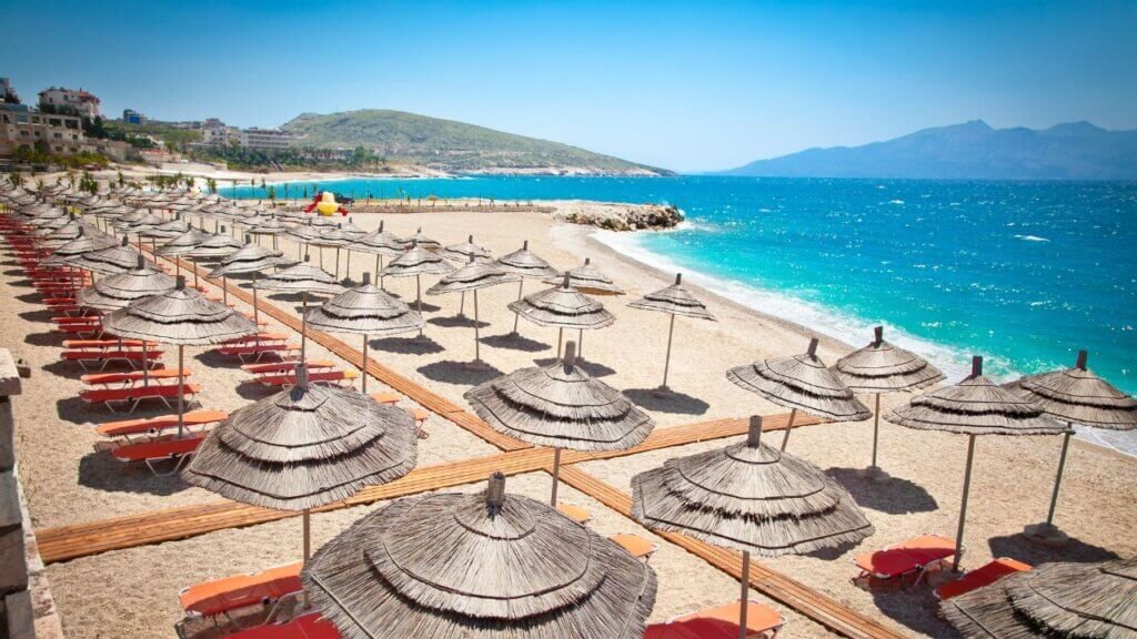 Rows of straw umbrellas and orange sun loungers on a sandy beach resort along the Albanian Riviera coastline