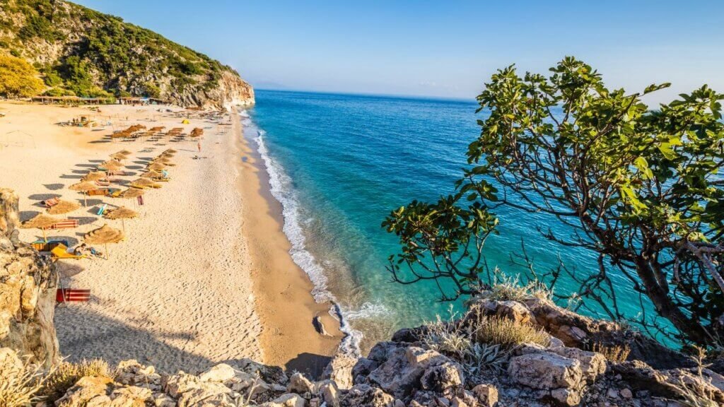 Aerial view from a cliff of a sandy beach lined with straw umbrellas and turquoise sea along the Albanian Riviera"
