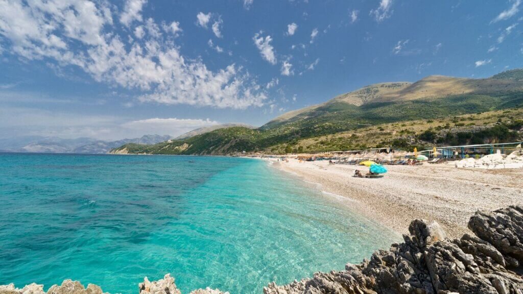 Pebble beach with turquoise and deep blue sea meeting the shore, backed by green mountains along the Albanian Riviera