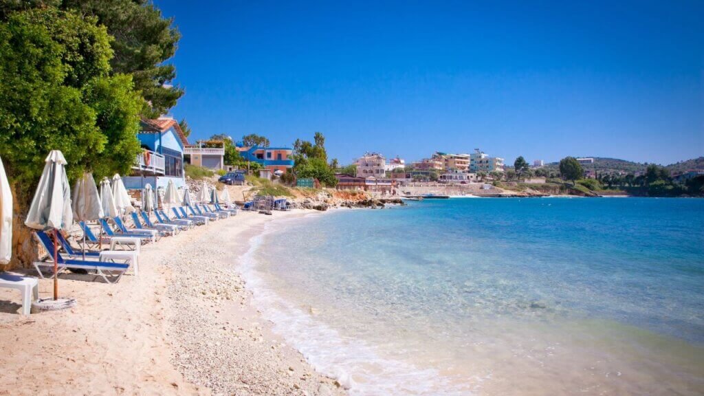 Beach loungers and umbrellas along a pebbly shoreline with colorful seaside houses and hotels in an Albanian coastal town