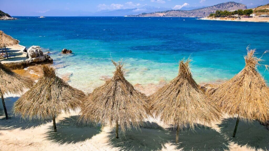 Aerial view from a cliff of a sandy beach lined with straw umbrellas and turquoise sea along the Albanian Riviera