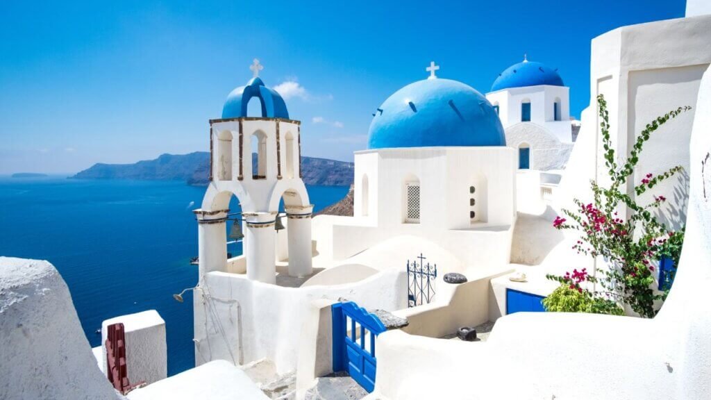 Classic white church bell tower and blue domes framed by bright pink bougainvillea flowers against the deep blue ocean in Santorini, Greece.