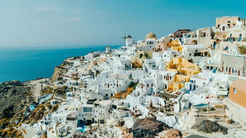 High-angle view of iconic blue-domed white churches in Oia overlooking the sparkling sunny Aegean Sea in Santorini.