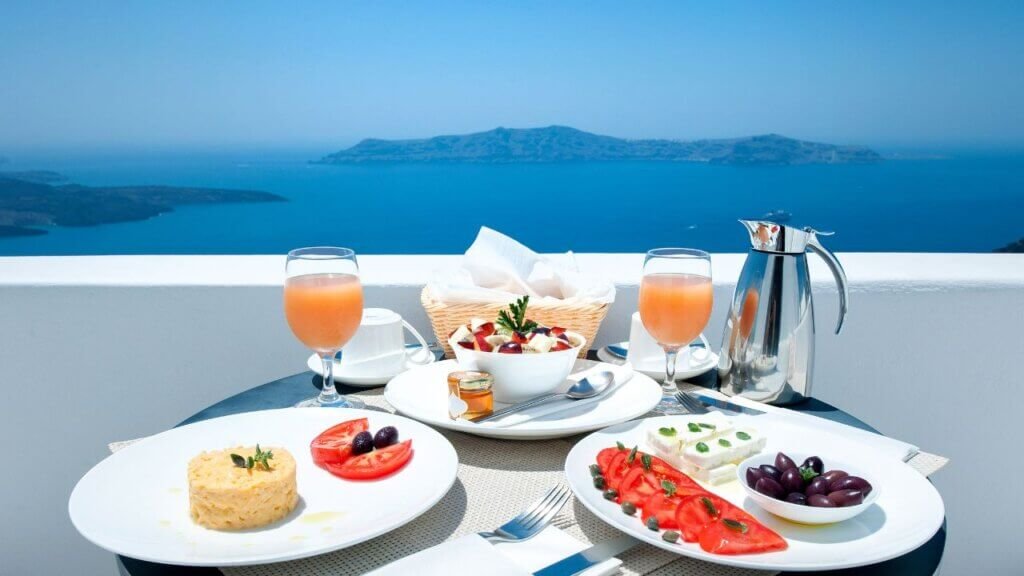 Mediterranean breakfast spread featuring fresh fruit, juice, cheese, and olives served on a balcony with a panoramic ocean view of Santorini.