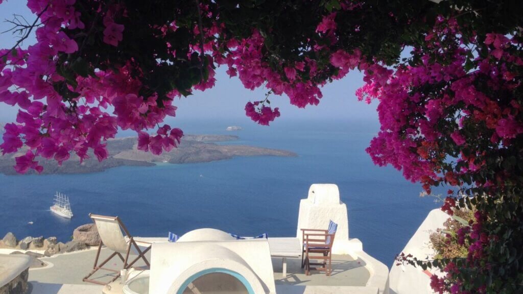 Outdoor private balcony in Santorini with lounge chairs and white architecture framed by blooming pink bougainvillea branches facing the ocean.